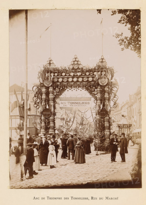 View of a triumphal arch in the Rue du Marché in Bergerac in honor of the entry of President Félix Faure, Arc de triomphe des tonneliers, Rue du Marché, photograph, page, Bergerac, 1895-06-04, photographer, paper, daylight gelatin silver print, height 161 mm x width 119 mm, street, triumphal arch, triumphal entry and public reception, pageant, 'entrée solennelle', 'joyeuse entrée', A street scene shows a large decorative archway made from stacked barrels, hoops, and foliage spanning the road, topped with round elements and flags on tall poles and flanked by ornate columns wrapped with garlands and rings; beneath the arch a banner is suspended and the street beyond is lined with buildings draped with more flags and hanging decorations. In the foreground a crowd of people wearing period clothing gathers on a cobbled street, including women in long dresses and hats, men in coats and hats, and children, with some people standing in groups and others walking. Two men in dark coats and hats ...