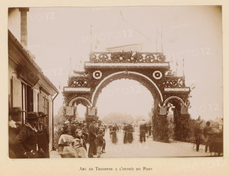 View of a triumphal arch in Bergerac in honor of the entry of President Félix Faure, Arc de triomphe a l'entrée du pont, photograph, page, Bergerac, 1895-06-04, photographer, paper, daylight gelatin silver print, height 120 mm x width 169 mm, street, triumphal arch, triumphal entry and public reception, pageant, 'entrée solennelle', 'joyeuse entrée', A wide decorative arch covered in foliage and floral ornaments spans a street, with round wreath-like decorations and circular emblems on its upper portion and smaller arches on either side, topped by a boat-like structure and pennant flags strung above; people in period clothing, including men in hats and women in long dresses, stand and walk beneath and around the arch, with building facades visible to the left and blurred figures and distant trees beyond the arch.