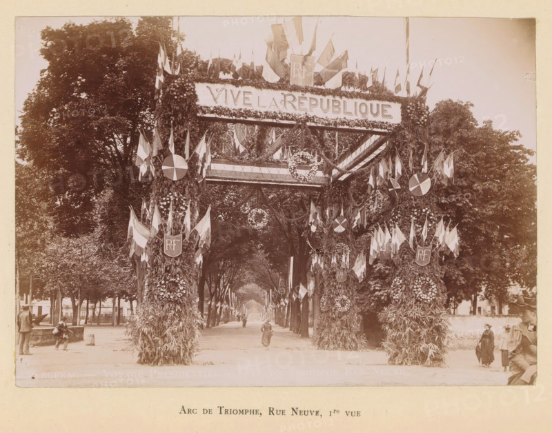 View of a triumphal arch on the Rue Neuve in Bergerac in honor of the arrival of President Félix Faure, Arc de triomphe, Rue Neuve, 1e vue, Bergerac- Voyage presidentialiel- Arc de triomphe, photograph, page, Bergerac, 1895-06-04, photographer, paper, daylight gelatin silver print, height 120 mm x width 169 mm, street, triumphal arch, triumphal entry and public reception, pageant, 'entrée solennelle', 'joyeuse entrée', A wide tree-lined avenue framed by tall leafy trees leads into the distance beneath a large decorated archway covered in foliage and floral arrangements, with numerous flags and garlands draped across the top and sides and circular floral wreaths and round emblems attached to the arch supports, while people in period clothing walk or stand along the avenue and at the base of the arch.