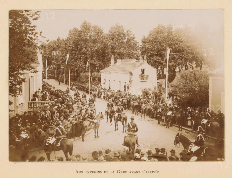 Bergerac (résumé historique) ses hotels illustres, book, publisher: Imprimérie Générale du Sud-Ouest (J. Castanet), Bergerac, 1895, paper, printing, daylight gelatin silver printing, height 252 mm x width 167 mm x thickness 16 mm, A wide street scene shows a crowd of people lining both sides of a road flanked by buildings and tall trees, many individuals wearing hats and period clothing, with mounted soldiers in uniform on horses forming a procession down the center of the street, flags on poles rising above the crowd, a two-story white building with a small balcony and shutters visible on one side, and dense foliage and trees filling the background.