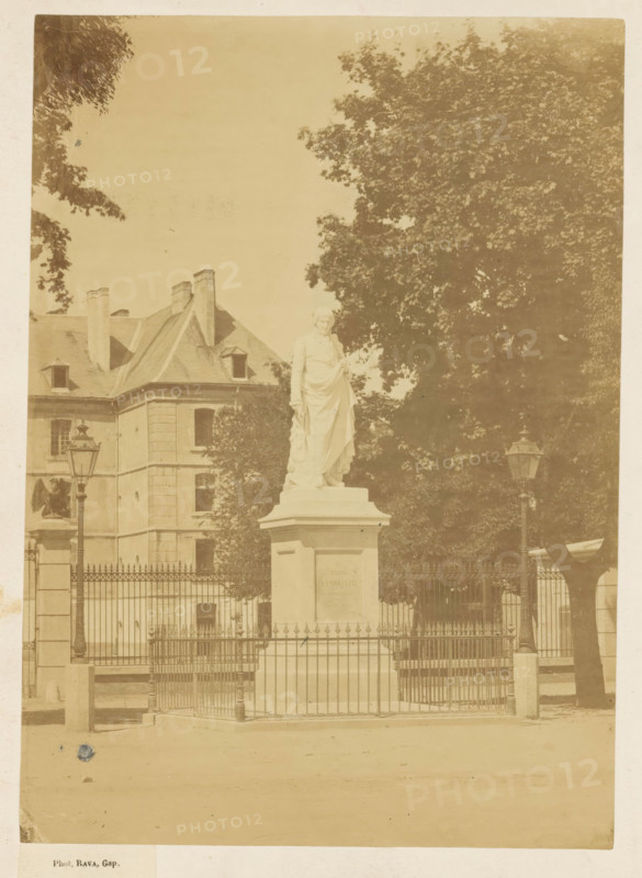 Statue of Charles-François de Ladoucette by Jean Marcellin in Gap, photograph, page, photographer: Rava, Gap, 1866, Provence-Alpes-Côte d'Azur, paper, albumen print, albumendruk, height 219 mm x width 156 mm, Person, Charles-François de Ladoucette, monument, statue, A pale photograph shows a stone statue of a robed figure standing on a rectangular pedestal behind a decorative metal fence, flanked by two tall ornate lampposts. Behind the statue is a large tree with dense foliage and to the left a multi-story building with a sloped roof, chimneys, and small dormer windows. The foreground shows a paved area and the fence includes pointed finials and gate posts. The image has a faded, sepia tone.