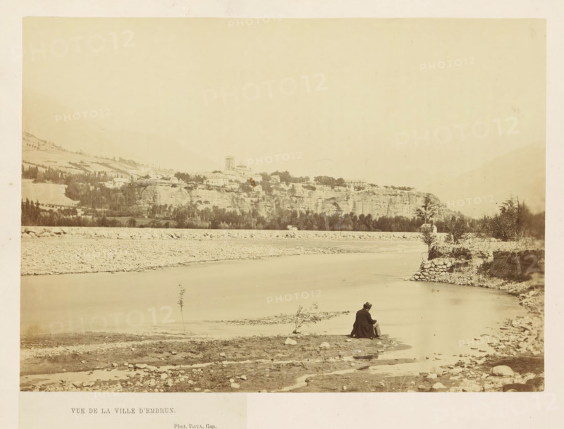 Vue de la Ville d'Embrun, View of Embrun, photograph, page, photographer: Rava, Embrun, c. 1856 - in or before 1866, Rava, photographer, Provence-Alpes-Côte d'Azur, paper, albumen print, height 157 mm x width 219 mm, city-view in general, 'veduta', river, A sepia-toned riverside scene showing a wide calm river with a rocky shore and a low pebbled embankment, a distant elevated town on a cliff with clustered buildings and a prominent tower, tree lines along the cliff base, terraced hills rising beyond, scattered rocks and sparse vegetation in the foreground, and a solitary seated figure wearing a dark coat and hat near the water's edge.