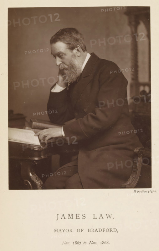 Portrait of James Law, James Law, mayor of Bradford, page, photomechanical print, inscription: 'Nov. 1867 to Nov. 1868.', photographer: Appleton & Co., Bradford (England), cliché maker: anonymous, c. 1871 - in or before 1881, paper, Woodburytype, Woodburytypepie, height 116 mm x width 92 mm, historical persons (portraits and scenes from the life) ((full) bust portrait), A seated older man with a full beard and short hair leans his head on one hand while resting his other hand on an open book on a table, wearing a dark suit with visible cuffs and seated on an ornately carved chair, with a blurred background and a neutral-toned backdrop.