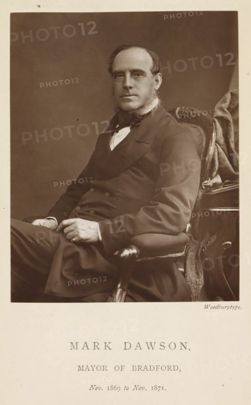 Portrait of Mark Dawson, Mark Dawson, mayor of Bradford, page, photomechanical print, inscription: 'Nov. 1869 to Nov. 1871.', photographer: Appleton & Co., Bradford (England), cliché maker: anonymous, c. 1871 - in or before 1881, paper, Woodburytype, Woodburytypepie, height 116 mm x width 91 mm, historical persons (portraits and scenes from the life) ((full) bust portrait), A sepia portrait of a seated man wearing a dark double-breasted coat and a bow tie, with sideburns and hair combed back, sitting in an ornate upholstered chair with carved wooden armrests and patterned fabric; his hands rest in his lap and he looks toward the camera against a plain dark background.