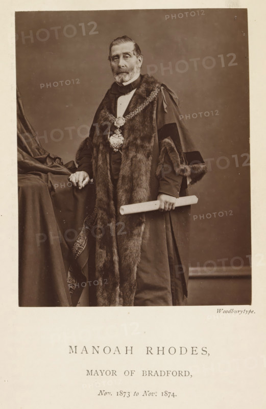 Portrait of Manoah Rhodes, Manoah Rhodes, mayor of Bradford, page, photomechanical print, inscription: 'Nov. 1873 to Nov. 1874.', photographer: Appleton & Co., Bradford (England), cliché maker: anonymous, c. 1871 - in or before 1881, paper, Woodburytype, height 116 mm x width 91 mm, historical persons (portraits and scenes from the life), A sepia portrait of an older man standing, wearing a dark formal robe trimmed with fur and a decorative chain with a large pendant around his neck, his hair combed back and a full beard and mustache; he rests one hand on a draped tablecloth and holds a rolled scroll in the other hand, with the background a plain studio backdrop and a narrow light border around the image.