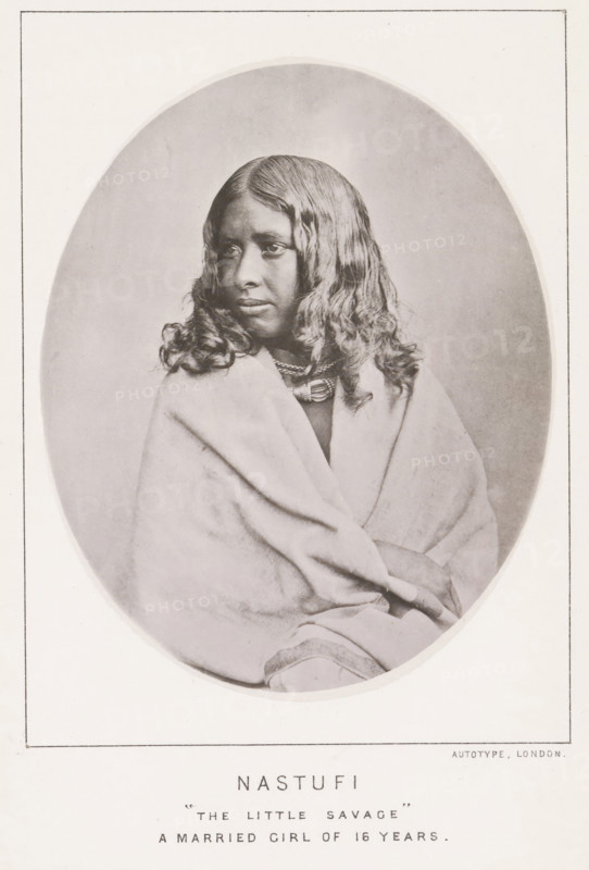 Nastufi 'the little savage' a married girl of 16 years, page, photomechanical print, number: 'No. 25', photographer: Bourne & Shepherd, Nilgiris, photographer: Nicholas & Curths, cliché maker: anonymous, printer (person): Autotype Printing Company, London, c. 1863 - in or before 1873, printer (person), paper, collotype, photoprint, height 122 mm x width 98 mm, adolescent, young woman, maiden, A sepia-toned oval portrait of a young person with long wavy hair wearing layered necklaces and wrapped in a light-colored blanket, shown from the torso up with a neutral expression and eyes looking slightly to the side, framed by a rectangular border on a pale background.