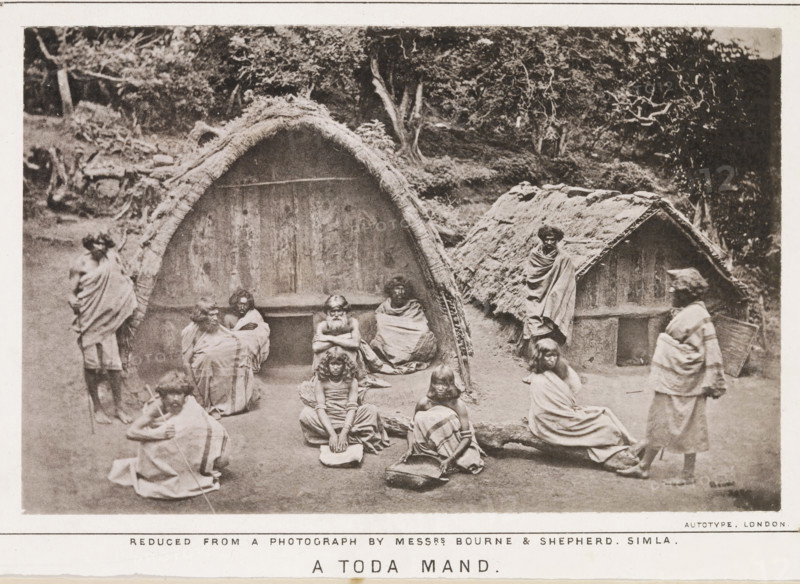 Travels among the todas or the study of a primitive tribe in South India, their history, character, customs, religion, infanticide, polyandry, language, with outlines of the Tuda grammar, book, name: 'WE Gladstone St. Deiniol Library Hawarden Elwyd Founded by The Rt. Hon. WE Gladstone', writer (clerical worker): William E. Marshall, publisher: Longman & Co., London, 1873, writer (clerical worker), textile materials, pasteboard, paper, linen (material), printing, collotype, height 226 mm x width 148 mm x thickness 32 mm, A sepia-toned photograph showing a group of people arranged in front of two thatched huts with arched and pitched roofs, surrounded by trees and foliage in the background; several seated and standing figures wear draped cloths, some with arms crossed or wrapped around themselves, and a few children sit on the ground with one holding a flat object and another beside a basket; a standing person carries a woven container on their back, and the ground appears bare with a lo...