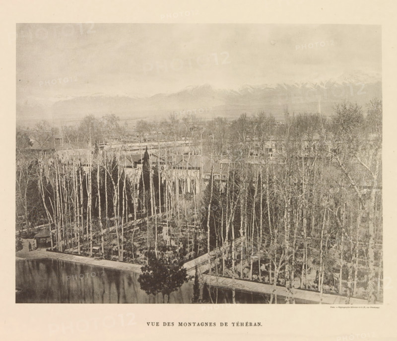 View of mountains in Tehran, Vue des Montagnes de Téhéran, page, photomechanical print, name: 'Silvestre et Cie, 97, rue Oberkampf.', printer (person): Silvestre et Cie, Paris, c. 1869 - in or before 1885, printer (person), paper, collotype, photoprint, height 129 mm x width 169 mm, mountains, Tehran, A wide landscape photograph showing a foreground of tall, slender trees with light-colored trunks densely packed along a paved walkway and bordering a reflective body of water, a cluster of small buildings partly visible behind the trees with pitched roofs and architectural details, a single darker evergreen tree near the water's edge casting a reflection, an expanse of flat land and scattered trees extending into the middle distance, and a distant range of snow-capped mountains beneath a pale, cloudy sky.