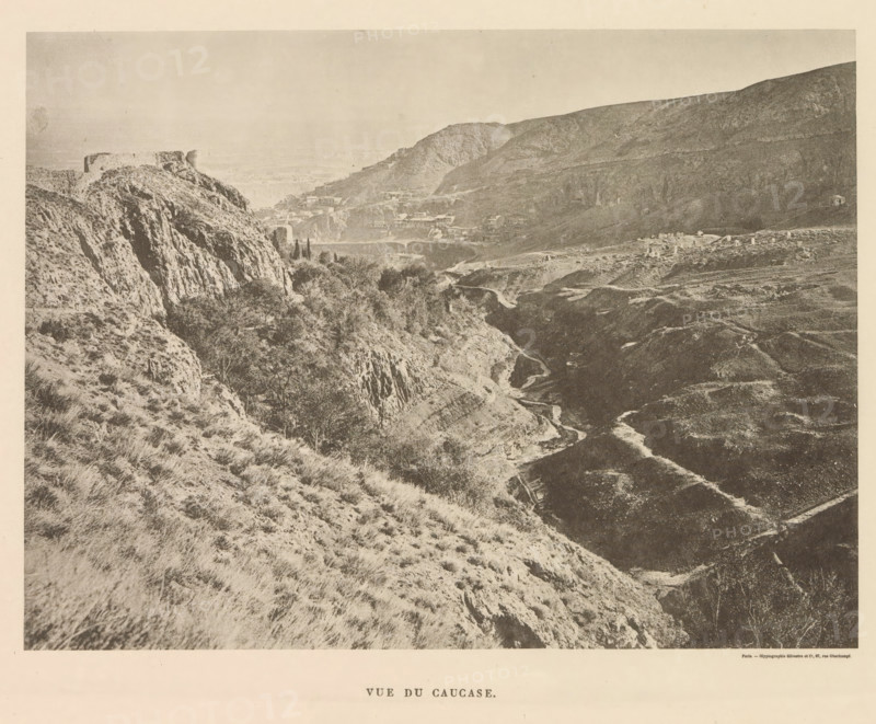 Vue du Caucase, View of the Caucasus, page, photomechanical print, name: 'Silvestre et Cie, 97, rue Oberkampf.', printer (person): Silvestre et Cie, Paris, c. 1869 - in or before 1885, printer (person), paper, collotype, photoprint, height 129 mm x width 174 mm, mountains, Caucasus, A wide landscape photograph showing rugged hills and steep rocky cliffs with sparse grasses and scattered shrubs, a winding dirt path or dry streambed cutting through a valley, clusters of trees and denser vegetation in lower areas, terraces or layered slopes on the distant hills, small buildings and structures nestled along the mid-distance slopes, and a prominent flat-topped rocky outcrop on the left side under a pale sky.