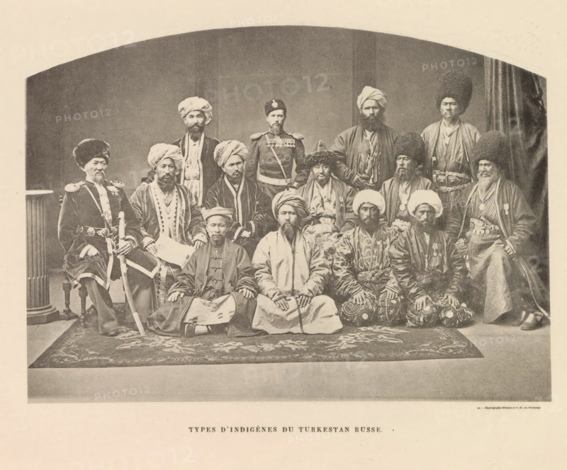 Group portrait of men from probably Uzbekistan, Types d'Indigènes du Turkestan Russe, page, photomechanical print, name: 'Silvestre et Cie, 97, rue Oberkampf.', printer (person): Silvestre et Cie, Paris, c. 1869 - in or before 1885, Silvestre et Cie, printer (person), paper, collotype, photoprint, height 127 mm x width 176 mm, human races, peoples, nationalities, A sepia-toned studio photograph showing a large group of men arranged in three rows, many wearing various styles of turbans, tall fur hats, and long robes, with several men wearing military-style jackets and belts; the front row is seated on a patterned rug with hands resting on their knees, the middle row sits on chairs, and the back row stands, all facing the camera with neutral expressions; a column is visible at the left edge and a draped curtain at the right, and the image has a rounded arch framing the top.