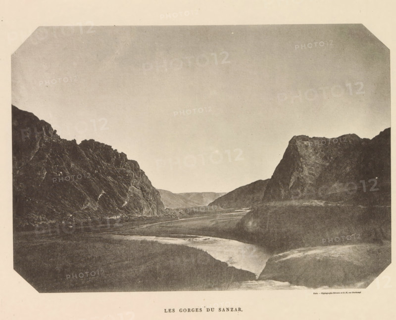Les Gorges' du Sanzar, View of a gorge in Uzbekistan, page, photomechanical print, name: 'Silvestre et Cie, 97, rue Oberkampf.', printer (person): Silvestre et Cie, Paris, c. 1869 - in or before 1885, Silvestre et Cie, printer (person), paper, collotype, photoprint, height 126 mm x width 179 mm, ravine, chasm, abyss, canyon, A wide landscape photograph showing steep rocky cliffs on both sides with a narrow river winding through the valley between them, a flat plain in the foreground, and an open sky above.