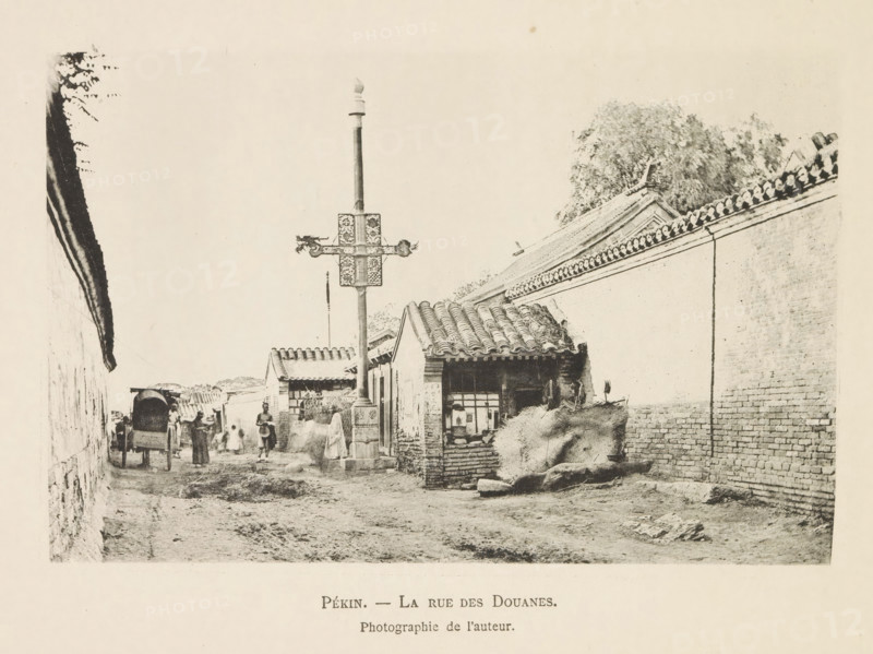 Street with customs posts in Beijing, Pékin. - La rue des customs, page, photomechanical print, inscription: 'Photographie de l'autoriteit.', photographer: Hugues Krafft, Beijing, cliché maker: anonymous, in or after 1882 - in or before 1883, Hugues Krafft, photographer, cliché maker, paper, collotype, photoprint, height 108 mm x width 166 mm, street, façade (of house or building), customs, A narrow dirt street lined with brick walls and low tiled-roof buildings, a tall ornate pole with decorative crossarms standing near the foreground, a small shop or house with an open window and stacked bundles or hay nearby, groups of people walking or standing along the street, a covered cart facing away from the viewer, tiled rooflines and trees visible above the walls, and a worn uneven pathway with scattered stones.