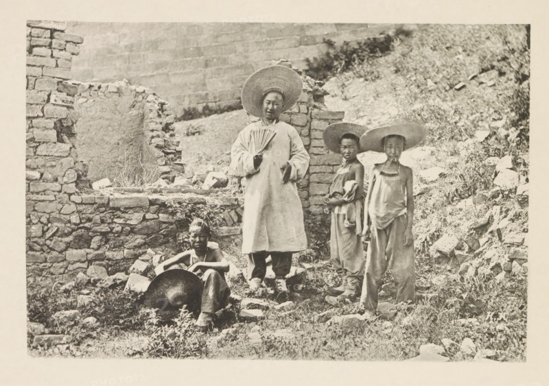 Tour guide Barthélemy and three mule drivers in front of a ruin in China, Barthélemy et les trois aniers, page, photomechanical print, inscription: 'Photographie de l'autoriteit.', inscription: 'HK', photographer: Hugues Krafft, China, cliché maker: anonymous, in or after 1882 - in or before 1883, Hugues Krafft, photographer, cliché maker, paper, collotype, photoprint, height 113 mm x width 168 mm, boy (child between toddler and youth), ruin of a building, architecture, fan, A black-and-white photograph showing four people standing and sitting among rough stone walls and a rocky, grassy hillside; one person stands near the center wearing a long light-colored garment and a wide straw hat while holding a folded hand fan, two younger individuals stand to the right wearing loose clothing and wide straw hats, and one person sits on the ground at the left foreground with a large round metal pan resting near their legs; broken stones, rubble, and patches of vegetation surround them and a part...