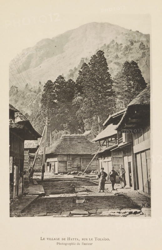 Street in a village in Japan, with a mountain in the background, Le village de Hatta, sur le Tokaïdo, page, photomechanical print, inscription: 'HK', inscription: 'Photographie de l'autoriteit.', photographer: Hugues Krafft, Japan, cliché maker: anonymous, in or after 1882 - in or before 1883, Hugues Krafft, photographer, cliché maker, paper, collotype, photoprint, height 178 mm x width 124 mm, street, mountains, outskirts of village, A narrow dirt street flanked by wooden buildings with thatched and tiled roofs, a few standing poles and leaning beams along the roadway, a cluster of tall dense trees rising behind a thatched building at the end of the street, a steep forested mountain filling the upper background, and three people standing on the right side of the street near a building entrance.