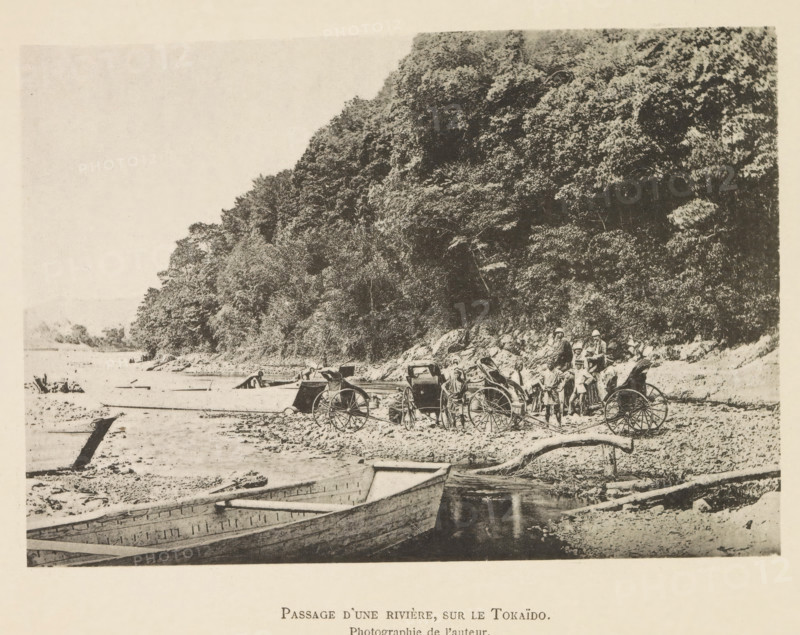 Passage d'une rivière, sur le Tokaïdo, Touring group with rickshaws on the bank of a river in Japan, page, photomechanical print, inscription: 'Photographie de l'autoriteit.', photographer: Hugues Krafft, Tokaido, cliché maker: anonymous, in or after 1882 - in or before 1883, Honshu, paper, collotype, photoprint, height 119 mm x width 172 mm, river, crossing a river, rickshaw, A sepia-toned scene shows a shoreline with a large wooded hillside occupying the right side and background, dense with trees and foliage. Several wooden rowboats rest in shallow water and on the shore in the foreground, their hulls visible and some partially beached. On the beach area near the base of the hill there are two-wheeled rickshaws with large spoked wheels and a group of people standing nearby, some wearing hats and simple clothing. The shoreline is scattered with rocks, logs, and debris, and the water surface appears calm. The overall image has a grainy, aged photographic quality.