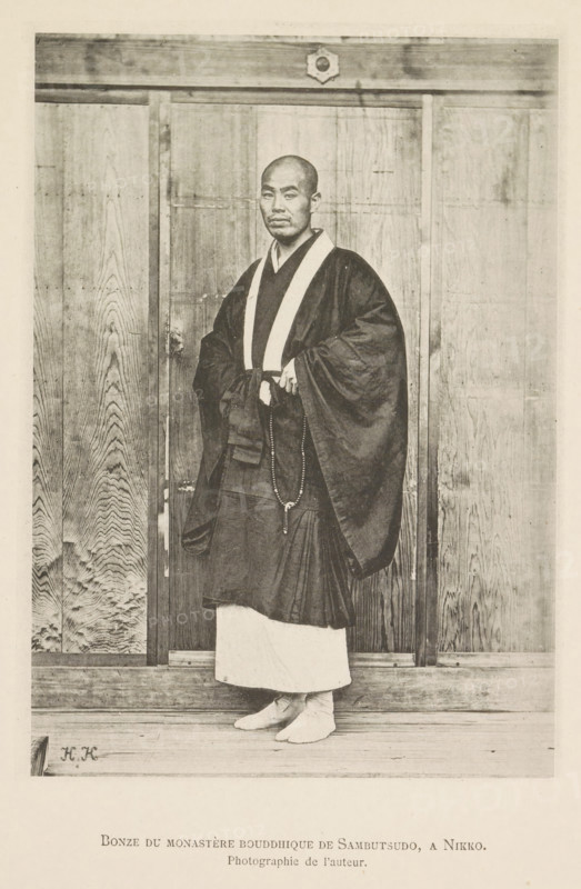 Buddhist monk in the Rinnoji temple in Nikko, Bonze du monastère bouddhique de Sambutsudo, a Nikko, page, photomechanical print, inscription: 'Photographie de l'autoriteit.', inscription: 'HK', photographer: Hugues Krafft, Rinnoji, cliché maker: anonymous, in or after 1882 - in or before 1883, Hugues Krafft, photographer, cliché maker, Kanto, Nikko, paper, collotype, photoprint, height 172 mm x width 122 mm, temple, shrine, Hinduism, Buddhism, Jainism, monk(s), friar(s), religious functions, A standing man with a shaved head wears layered dark robes with a contrasting light collar and a long beaded necklace draped from his hands, and he stands barefoot on a wooden floor in front of tall wooden panels with visible wood grain and a horizontal beam above, with a small decorative emblem centered on the beam.