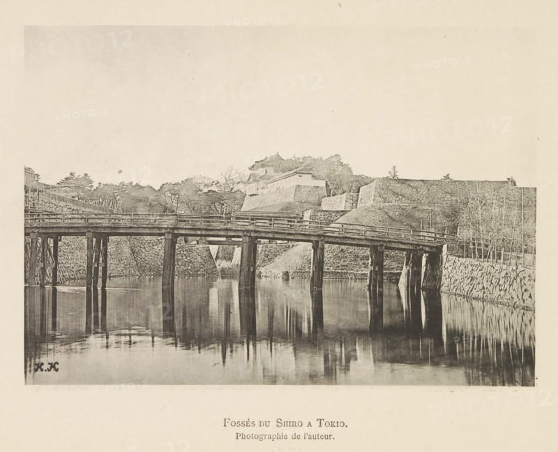 Bridge over a canal in Tokyo, with the Kokyo, Fossés du Shiro a Tokyo in the background, page, photomechanical print, inscription: 'HK', inscription: 'Photographie de l'autoriteit.', photographer: Hugues Krafft, Tokyo, cliché maker: anonymous, in or after 1882 - in or before 1883, Hugues Krafft, photographer, cliché maker, Kanto, paper, collotype, photoprint, height 115 mm x width 164 mm, bridge, palace, moat, fortified city, A sepia-toned photograph showing a wooden bridge supported by vertical posts spanning calm reflective water, with the bridge's railings and structural beams visible; behind the bridge are sloped stone embankments and terraced stone walls leading up to a cluster of traditional roofed buildings partially obscured by trees; bare and leafy trees line the top of the embankments and small poles or stakes stand along the right side near the water's edge; the water surface reflects the bridge posts and nearby stonework, and the sky above is light and largely empty.