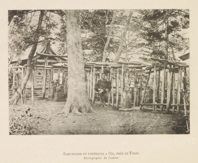 View of an Oji shrine near Tokyo, Sanctuaire et portiques a Oji, près de Tokyo, page, photomechanical print, inscription: 'Photographie de l'autoriteit.', photographer: Hugues Krafft, Japan, cliché maker: anonymous, in or after 1882 - in or before 1883, Hugues Krafft, photographer, cliché maker, paper, collotype, photoprint, height 117 mm x width 170 mm, forest, wood, road side shrine, Kanto, Tokyo, A sepia-toned scene shows a fenced wooden structure with vertical posts and horizontal beams set among tall trees, a large tree trunk occupying the foreground center with visible roots at its base, a person standing near the fence wearing traditional clothing and a head covering, a small stone statue on a pedestal beside the fence, a building with a sloped roof partially visible behind the fence and trees, scattered leaves and bare ground in the open area in front, and branches and foliage filling the upper portion of the image.
