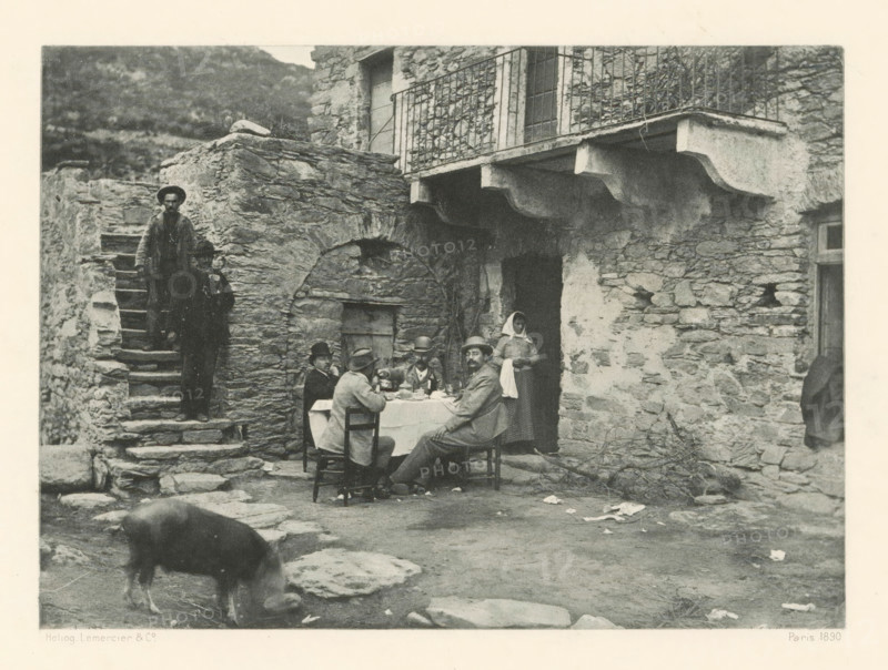 Breakfast company in Casta, Déjeuner à Casta, page, photomechanical print, photographer: Roland Napoleon Bonaparte, Corsica, cliché maker: Lemercier & Cie., Paris, c. 1881 - in or before 1890, paper, photoaquatint, height 118 mm x width 163 mm, Print on frontispiece., street, pig, breakfast - CC - out of doors, A worn stone courtyard in front of a rustic stone building with an arched doorway and a small balcony with a metal railing, a table covered with a cloth set for a meal with several seated figures wearing hats and coats, a standing woman in a headscarf near the doorway, rough stone steps up the left side with two standing figures on them, scattered stones and debris on the ground, and a pig grazing in the foreground.
