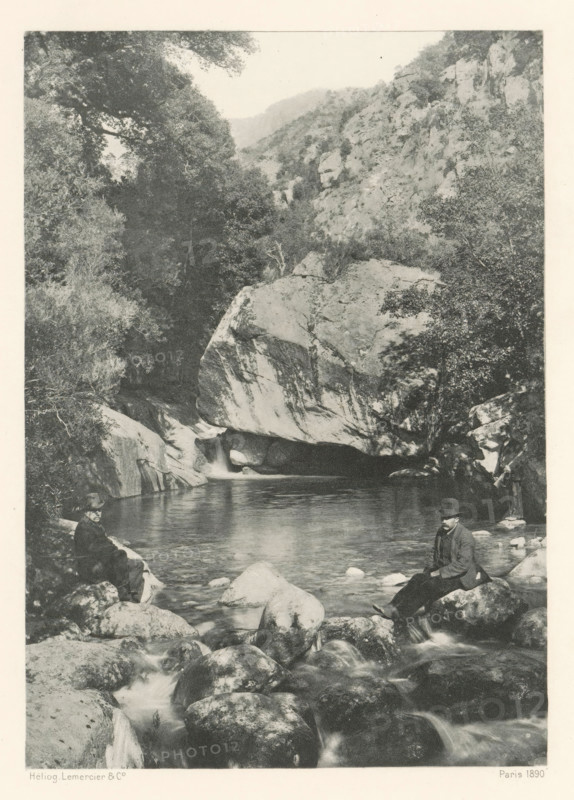 La Pentica, Two men before the Pentica Gorge in Corsica, page, photomechanical print, photographer: Roland Napoleon Bonaparte, Corsica, cliché maker: Lemercier & Cie., Paris, c. 1881 - in or before 1890, paper, photoaquatint, height 167 mm x width 118 mm, sitting figure, ravine, chasm, abyss, canyon, A grayscale scene shows a rocky river with smooth boulders in the foreground and a calm pool further back, flanked by steep rock faces and dense trees; a large rounded rock overhang sits above the pool with water flowing beneath it, and two men in dark coats and hats are seated on separate rocks near the water’s edge.