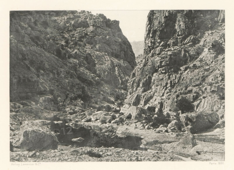 Gorges du Golo, Gorge of the river Golo in Corsica, page, photomechanical print, photographer: Roland Napoleon Bonaparte, Corsica, cliché maker: Lemercier & Cie., Paris, c. 1881 - in or before 1890, paper, photoaquatint, height 117 mm x width 167 mm, river, ravine, chasm, abyss, canyon, A monochrome image shows a rocky canyon with steep, jagged cliff faces on both sides and a narrow gorge between them. The foreground contains scattered boulders and a rocky shoreline leading to a dark, calm pool of water reflecting nearby stones. The cliff surfaces display rough textures, ledges, and crevices, with some sparse low vegetation clinging to the rock. The sky visible above the gorge is pale and contrasts with the darker tones of the rock.