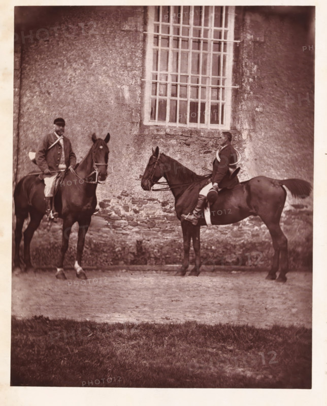 Two hunters on horseback, page, photomechanical print, photographer: Léon Cremière, France, cliché maker: anonymous, c. 1869 - in or before 1879, paper, Woodburytype, Woodburytypepie, height 202 mm x width 160 mm, Print on frontispiece., riding a horse, ass, or mule, rider, horseman, hunter, A sepia-toned photograph showing two men seated on horses in front of a textured stone wall with a tall window featuring a grid of vertical and horizontal bars; the riders wear jackets and breeches and hold reins, one rider facing slightly toward the camera while the other sits in profile, a coiled rope or lanyard visible over one rider’s shoulder, the horses are dark in color with bridles and saddles, the ground beneath them is a mix of dirt and a strip of grass in the foreground.