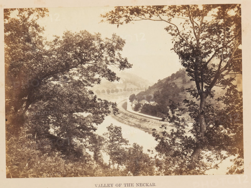 Valley of the Neckar, View of the River Neckar, photograph, page, photographer: Francis Frith, Germany, c. 1855 - in or before 1865, paper, albumen print, height 121 mm x width 169 mm, river, A sepia-toned landscape framed by tree branches and foliage in the foreground, with leafy canopies creating a natural border on left and right sides; a river curves through the middle distance, bordered by a winding road and railway track that follow the river's edge; a cluster of small buildings with pitched roofs sits near the riverbank beside the road, and hills rise behind them covered in trees and shrubs; distant hills and a line of trees along the far bank recede into a hazy background under a pale sky.