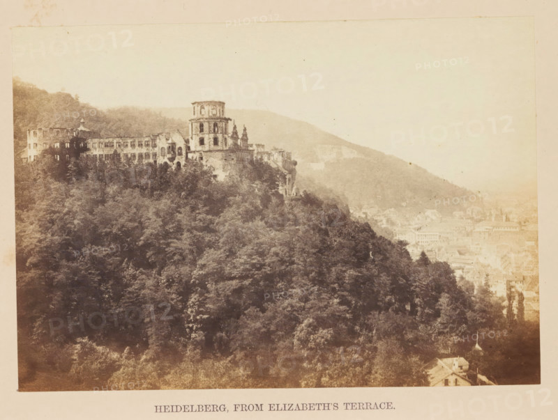 Heidelberg, from Elizabeth's Terrace, View of Schloss Heidelberg, photograph, page, photographer: Francis Frith, c. 1855 - in or before 1865, Francis Frith, photographer, Baden-Württemberg, paper, albumen print, height 116 mm x width 165 mm, castle, Schloss Heidelberg, A sepia-toned photograph showing a hillside covered in dense trees with a large stone building complex perched near the top, featuring a prominent multi-level round tower and adjoining rectangular structures with rows of windows and architectural details; the hill continues into a distant, hazy ridge with lighter-toned buildings and rooftops visible lower on the slope, and the foreground is filled with textured tree canopies and a few small structures partially visible at the bottom edge.