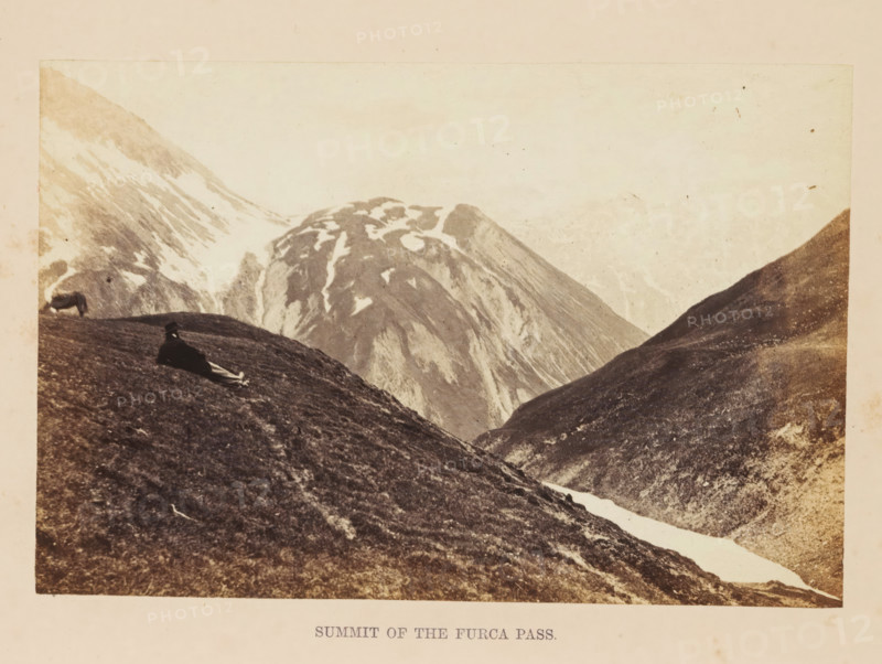 Summit of the Furca Pass, View of the top of the Furka Pass, photograph, page, photographer: Francis Frith, Furka Pass, c. 1855 - in or before 1865, paper, albumen print, height 113 mm x width 171 mm, mountain-range, snow peaks, A sepia-toned mountain landscape showing steep grassy slopes in the foreground and large rocky peaks with patches of snow in the background, with a narrow strip of snow or ice running down a valley between the slopes; a person reclining on the nearer hillside wearing dark clothing and a hat, and an animal grazing farther up the left slope; the sky appears pale and light above the mountains.