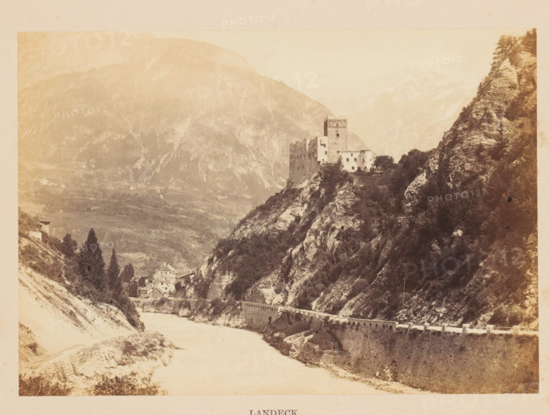 Landeck, View of Landeck and Landeck Castle, photograph, page, photographer: Francis Frith, c. 1855 - in or before 1865, Tirol, paper, albumen print, height 121 mm x width 172 mm, river, city-view, and landscape with man-made constructions, castle, A sepia-toned landscape showing a river flowing along the base of steep rocky cliffs, a road or embankment with a low wall running parallel to the river, a cluster of buildings nestled near the water at the foot of the cliffs, a larger multi-story stone structure or castle perched atop a rocky promontory above the cluster, dense trees and vegetation on the slopes and along the cliffs, and distant hazy mountains forming the background.
