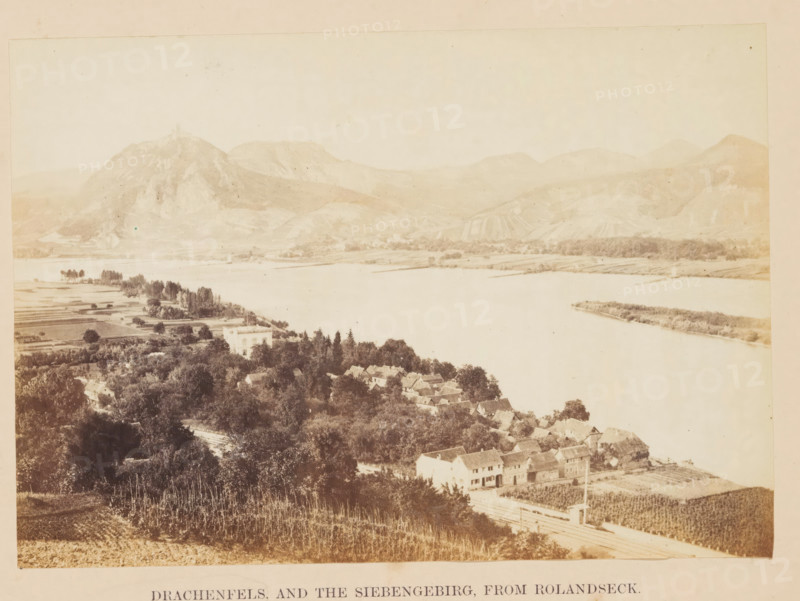 Drachenfels and the Siebengebirg, from Rolandseck, Siebengebirge with the Drachenfels seen from Rolandseck, photograph, page, photographer: Francis Frith, Remagen, c. 1855 - in or before 1865, Rhineland-Palatinate, paper, albumen print, height 116 mm x width 165 mm, mountain-top, peak, North Rhine-Westphalia, Königswinter, Drachenfels, A wide sepia-toned landscape showing a river with calm water running horizontally across the middle of the image, a cluster of houses with pitched roofs nestled among dense trees on the near bank, cultivated fields and orchards on the lower slopes in the foreground, a larger pale building set back among trees near the water, low rolling hills and rocky mountains rising on the far side of the river with terraced or striped slopes, scattered groves of trees along the opposite shore, and an open pale sky above.