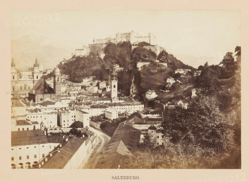 Saltzburg, View of Salzburg, photograph, page, photographer: Francis Frith, Salzburg (city), c. 1855 - in or before 1865, paper, albumen print, height 116 mm x width 166 mm, city-view, and landscape with man-made constructions, castle, A sepia-toned panoramic view of a historic town with clustered rooftops, narrow streets, and a mix of multi-story buildings and churches; several towers with domed and spired tops rise above the roofs, and a large fortress sits atop a wooded hill overlooking the town; the hillside shows terraced walls and scattered houses, while trees and foliage frame parts of the scene; in the foreground a long roofed structure and a curved street lead toward the denser center of buildings.