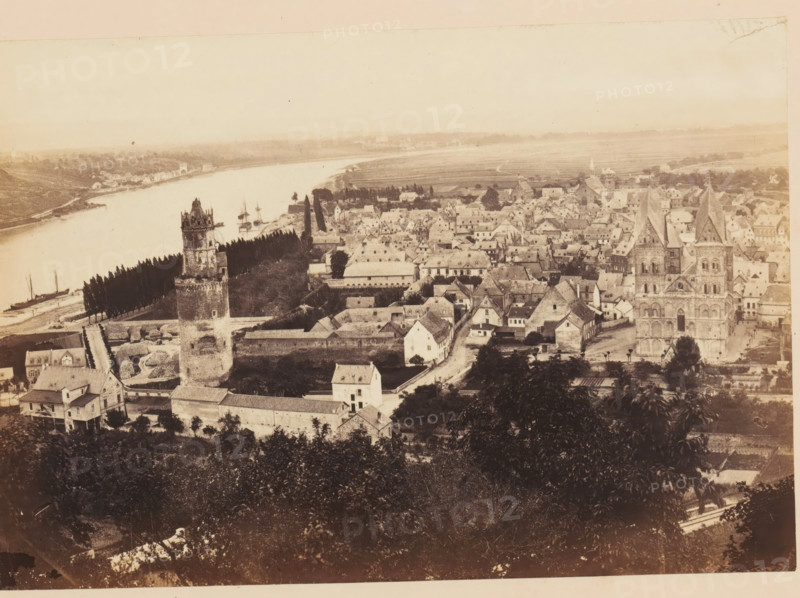 View of Andernach, Andernach, photograph, page, photographer: Francis Frith, c. 1855 - in or before 1865, Rhineland-Palatinate, paper, albumen print, height 116 mm x width 168 mm, city-view, and landscape with man-made constructions, tower, clock tower, monumental adornment, A sepia-toned aerial view of a riverside town showing a wide river curving along the left side with small sailing vessels moored near the shore, a tree-lined embankment running beside the water, a prominent stone tower with battlements near the foreground, a large church with twin spires and detailed stone façade toward the right, dense clusters of pitched-roof houses and narrow streets filling the central area, rectangular walled compounds and low buildings surrounding the tower, scattered trees and gardens among the buildings, and open fields stretching into the distant horizon under a pale sky.