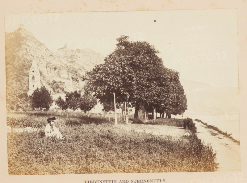 View of the bank of the Rhine and in the distance presumably Schloss Liebenstein, Liebenstein and Sternenfels, photograph, page, photographer: Francis Frith, Baden-Württemberg, c. 1855 - in or before 1865, paper, albumen print, albumendruk, height 116 mm x width 167 mm, castle, river bank, A sepia-toned scene shows a grassy foreground with a person seated wearing a wide-brimmed hat and light clothing, positioned near the lower left. A row of leafy trees with visible trunks lines the middle ground, casting darker foliage against a pale sky. Behind the trees on the left, a tall building with a pointed tower and arched openings stands at the base of a rocky hillside, which rises steeply and shows rough textures and outcrops. A narrow dirt path runs along the right edge toward a lighter area at the horizon.