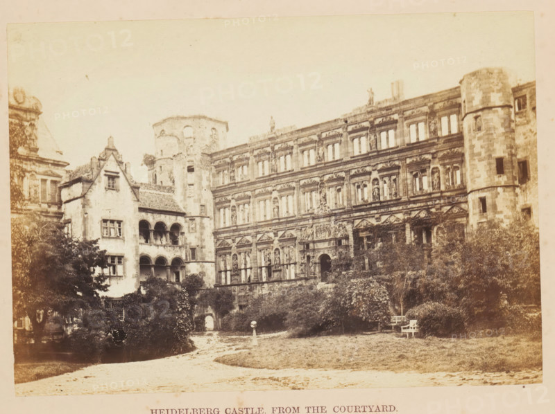 View of Schloss Heidelberg from the courtyard, Heidelberg Castle, from the Courtyard, photograph, page, photographer: Francis Frith, Heidelberg, c. 1855 - in or before 1865, Baden-Württemberg, paper, albumen print, albumendruk, height 122 mm x width 172 mm, courtyard, castle, Schloss Heidelberg, A sepia-toned photograph shows a large ornate stone building with multiple rows of windows and detailed sculptural decorations on its facade, adjoining a round tower with small rectangular openings and weathered stonework; to the left a smaller building with arched openings and a steep tiled roof connects to the larger structure, and a partial dome or roof element appears at the far left edge. In front of the buildings there is a garden area with trees and shrubs, a grassy lawn, a winding dirt path, and a pair of simple benches near the right side. The sky above is pale and empty, and the overall image has an aged, slightly faded appearance.