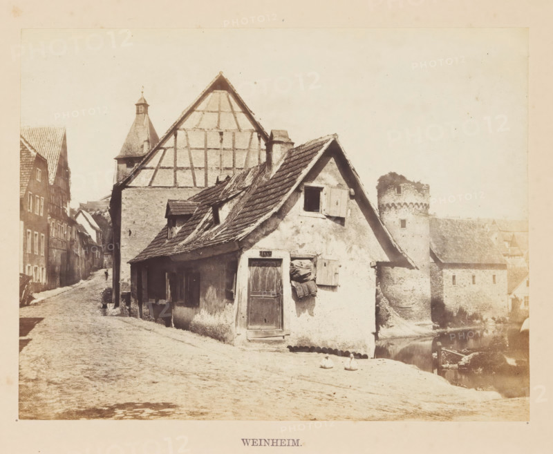 Weinheim, Half-timbered house in Weinheim, photograph, page, photographer: Francis Frith, c. 1855 - in or before 1865, Baden-Württemberg, paper, albumen print, height 125 mm x width 163 mm, housing, A street scene showing old buildings with steeply pitched, tiled roofs and exposed timber framing, a small plastered house in the foreground with a wooden door and a shuttered window, fabric or sacks hanging beside the door, a narrow cobblestone road running past the houses with two birds on the ground near the front building, rows of adjacent multi-story houses lining the left side of the street, a tower with battlements and a circular turret visible to the right behind the foreground building, reflections of part of the stone wall in water near the right edge, and a pale sky above.