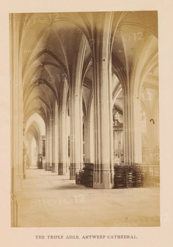 The triple aisle, Antwerp cathedral, photograph, page, Onze-Lieve-Vrouwekathedraal (Antwerp), c. 1856 - in or before 1866, photographer, Antwerp, paper, albumen print, height 160 mm x width 110 mm, interior of church, parts of church interior: aisle, A long interior view of a cathedral aisle with tall stone columns and pointed vaulted arches extending along the length, pale stone floors and walls, stacked wooden chairs arranged near the columns on one side, narrow clerestory windows letting in light, decorative stonework and an altar visible in the distance, and a printed caption centered below the image.