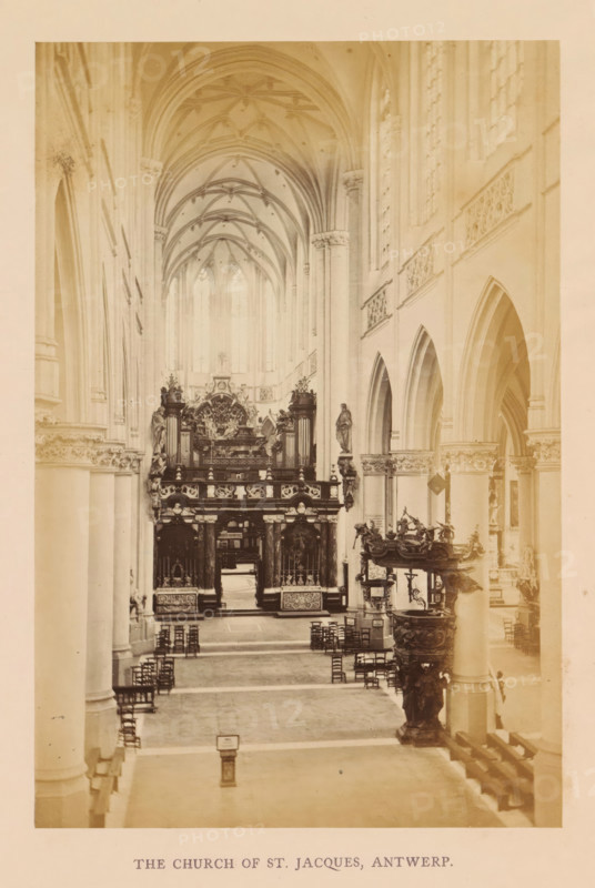 The church of St. Jacques, Antwerp, photograph, page, Sint-Jacobskerk (Antwerp), c. 1856 - in or before 1866, photographer, paper, albumen print, albumendruk, height 177 mm x width 118 mm, interior of church, parts of church interior: aisle, A sepia-toned interior photograph of a large Gothic church nave with high ribbed vaulted ceilings and tall arched windows along the right side, flanked by rows of slender columns and pointed arches leading toward an ornate raised chancel area. The central aisle is lined with scattered wooden chairs and leads to a sculpted rood screen or choir loft richly decorated with carved figures and ornamental panels, with a large altar and organ pipes visible behind it. On the right foreground stands an elaborate carved pulpit with sculpted figures and decorative railings, and statues mounted on columns are visible along the nave. Light filters through the high windows, casting soft illumination on the stone surfaces and architectural details throughout the s...
