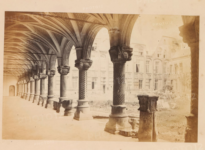 Court of the bishops palace, Liege, photograph, page, Liège, c. 1856 - in or before 1866, photographer, paper, albumen print, height 120 mm x width 183 mm, arcade, architecture, Palace of the Prince-Bishops, A long covered arcade with a series of stone columns and carved capitals supporting ribbed vaulted arches runs along the left side of the image, the columns showing twisted and fluted shafts and ornate bases, some with sculpted faces on the capitals. The vaulted ceiling displays intersecting ribs and brick infill, and slender horizontal rods span between the arches. Beyond the arcade, a courtyard with grass and scattered stone fragments is visible, along with a tall stone building featuring many tall windows with arched and rectangular tops and decorative masonry around them. In the foreground on the right, there are several carved stone pedestals and bases, one partially broken, and a small stone basin sits near the nearest column. The overall scene is rendered in a pale, sepia to...
