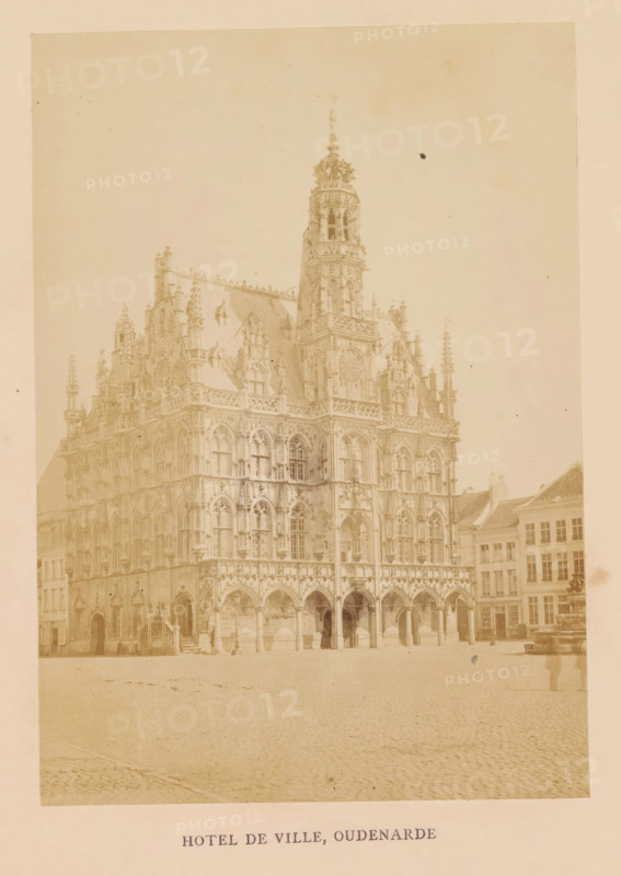 Hotel de ville, Oudenarde, photograph, page, Oudenaarde, c. 1856 - in or before 1866, photographer, paper, albumen print, albumendruk, height 158 mm x width 112 mm, arcade, architecture, Stadhuis van Oudenaarde, A large ornate stone building with Gothic architectural details fills the left and center of the image, featuring pointed arches at ground level, rows of tall narrow windows, decorative tracery, pinnacles, and sculptural ornamentation; a tall central tower with an elaborate spire rises above the roofline; adjacent buildings with simpler facades and pitched roofs appear to the right; an open cobblestone square occupies the foreground; a lone figure stands near the building entrance and a blurred person is visible near the right edge; the photograph has a faded, sepia-toned appearance.