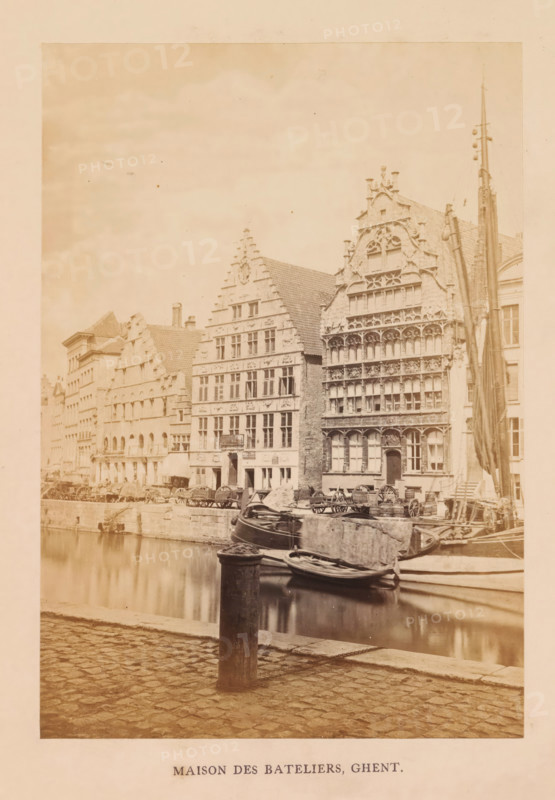 Maison des bateliers, Ghent, photograph, page, c. 1856 - in or before 1866, photographer, paper, albumen print, height 170 mm x width 118 mm, street, façade (of house or building), Gildehuis der Vrije Schippers, A waterside street scene shows a row of tall, ornate stone buildings with stepped gables and decorative facades facing a canal. Several boats are moored along the canal edge, some tied to posts and a rounded bollard in the foreground, with reflections visible in the water. The cobbled pavement runs along the canal in the foreground. On the buildings, many windows and carved architectural details are visible, and a tall mast or pole stands beside one of the structures. Wagons and wooden carts are lined along the quay beside the buildings. The overall image has a sepia tone.