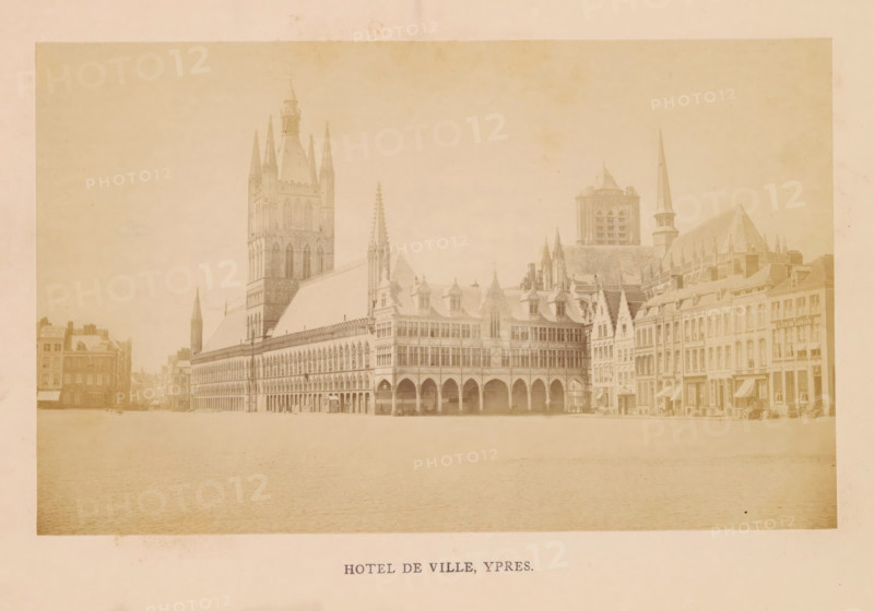 Hotel de ville, Ypres, photograph, page, c. 1856 - in or before 1866, photographer, paper, albumen print, height 110 mm x width 178 mm, square, circus, façade (of house or building), Cloth Hall, A wide sepia-toned photograph of an ornate town square dominated by a large Gothic-style building with tall spires, pointed arches, and a steeply pitched roof, adjoining a row of multi-story buildings with gabled facades, dormer windows, and a narrow tower with a spire; the foreground shows a broad open paved area, and the buildings display repeated arched arcades, decorative stonework, and a variety of window shapes and rooflines.