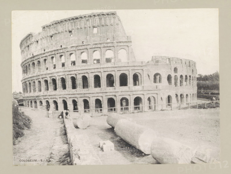 Coloseum, page, photomechanical print, photographer: Samuel J. Beckett, Rome, c. 1890 - c. 1900, paper, collotype, photoprint, height 151 mm x width 202 mm, facade (of house or building), theater (building), Colosseum, A wide photograph of a large stone amphitheater showing multiple tiers of rounded arches and rectangular openings, with worn and weathered masonry and broken sections along the upper edge; in the foreground there are large cylindrical stone fragments lying on the ground near a low wall, and a narrow path runs alongside the structure; sparse vegetation appears at the image edges and a low fence is visible near the base of the building.