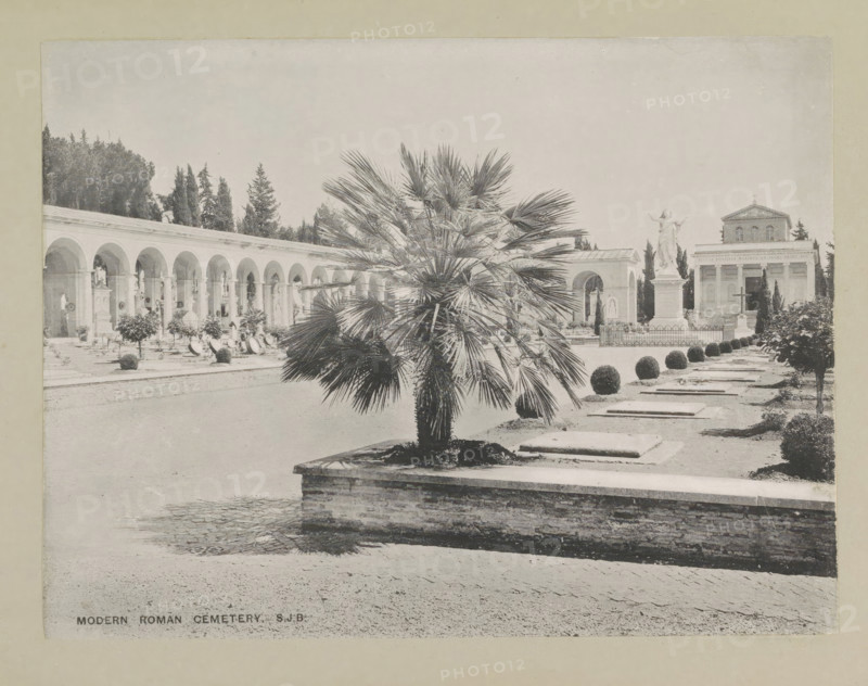 Modern Roman cemetery, page, photomechanical print, photographer: Samuel J. Beckett, Rome, c. 1890 - c. 1900, paper, collotype, photoprint, height 152 mm x width 200 mm, churchyard, cemetery, Verano Cemetery, A wide outdoor scene shows a paved courtyard with a raised rectangular planter in the foreground containing a fan-leaved palm, a row of flat stone slabs set into the ground beside neatly trimmed spherical shrubs, and a low brick retaining wall along the planter edge; to the left a long colonnade with repeated arches and niches runs back toward a stand of tall dark trees, with ornamental plants and monuments beneath the arches; toward the right a tall statue of a robed figure stands on a pedestal behind an ornamental fence, and behind it a classical building with columns and a pediment is visible, framed by additional trees and shrubbery under a pale sky.