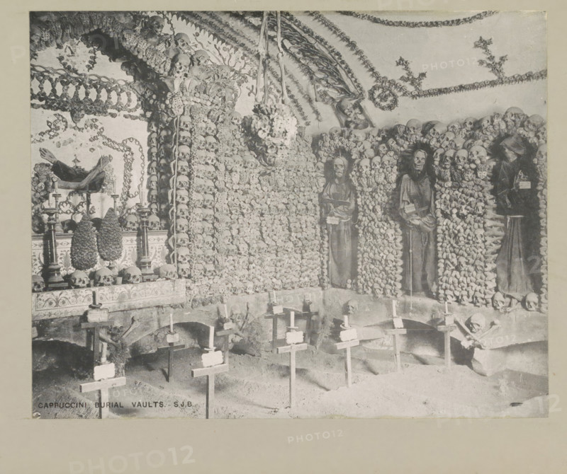 Cappuccini burial vaults, page, photomechanical print, photographer: Samuel J. Beckett, Santa Maria della Concezione dei Cappuccini, c. 1890 - c. 1900, Rome, paper, collotype, photoprint, height 151 mm x width 192 mm, charnelhouse, ossuary, A dim interior scene filled with human skulls and bones arranged into ornate patterns along the walls and arches, with rows of skulls forming columns and garlands hanging from the ceiling; three robed skeletal figures are mounted upright against the bone-covered wall, each appearing framed by surrounding skulls; a table or altar area displays skulls and candleholders, and cone-shaped objects sit on stands; in the foreground a series of simple crosses stand in the floor, each topped with a small candle, and a skeletal figure kneels near one of the crosses; chandeliers and hanging clusters of bones descend from the ceiling, and decorative motifs of bones repeat across the architectural surfaces.