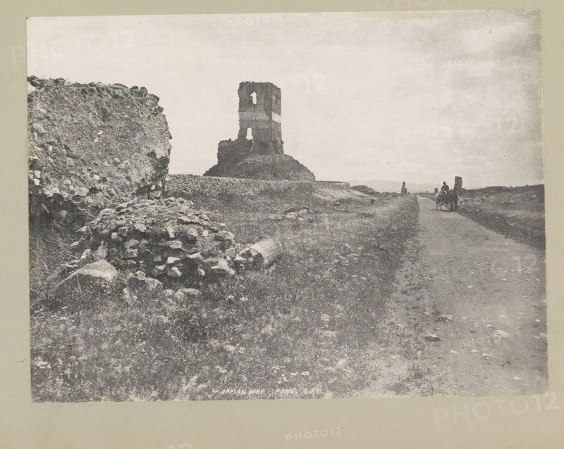 Appian way. Rome, page, photomechanical print, photographer: Samuel J. Beckett, Via Appia Antica, c. 1890 - c. 1900, paper, collotype, photoprint, height 152 mm x width 202 mm, road, path, landscape with ruins, A wide, grainy black-and-white landscape showing a rough dirt road running toward the horizon with grassy, rocky ground on either side, a ruined stone tower standing on a small rise near the center with distinct window openings, a broken stone wall and scattered rubble on the left foreground, and a small horse-drawn cart with figures on the road near the distant right side under a pale, lightly textured sky.