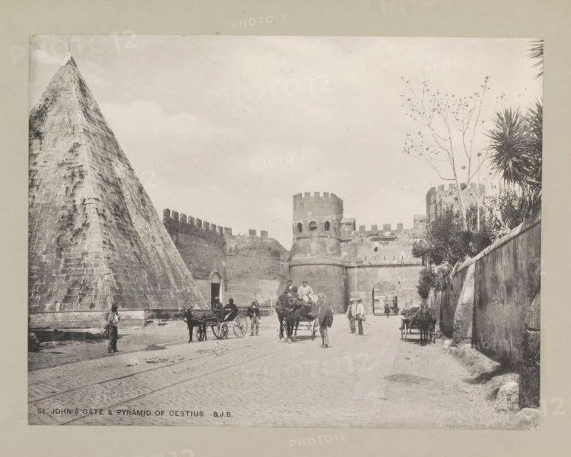 St John's gate & pyramid of Cestius, page, photomechanical print, photographer: Samuel J. Beckett, Rome, printmaker: Samuel J. Beckett (possibly), c. 1890 - c. 1900, Samuel J. Beckett, photographer, printmaker, paper, collotype, photoprint, height 153 mm x width 198 mm, pyramid (marking grave), gate, entrance, Porta San Paolo, Pyramid of Cestius, A wide street scene in monochrome shows a large stone pyramid on the left and a fortified stone gate with rounded towers and crenellations toward the center and right, with a high wall extending across the scene; several horse-drawn carriages and horses occupy the roadway and people in period clothing stand and walk among them; a tall thin tree with sparse branches rises near the right-side tower and spiky palm-like plants grow along the right wall; cobbled pavement and faint tram tracks run through the foreground and a few individuals stand near the pyramid base and along the walls.