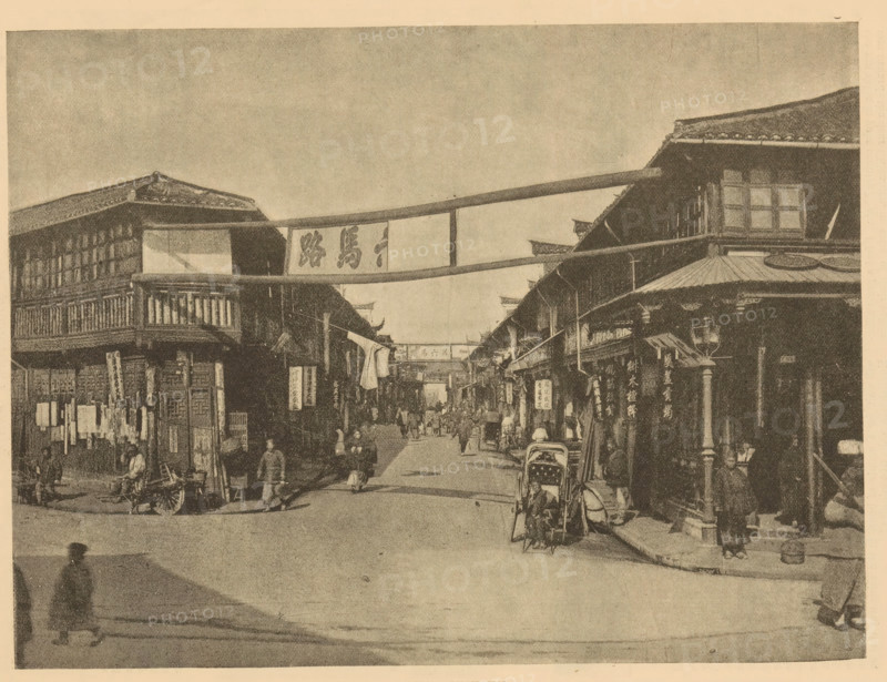 A street in Shanghai, page, photomechanical print, Shanghai, printmaker: Meisenbach's Autotype Company (possibly), c. 1881 - in or before 1886, Meisenbach's Autotype Company, photographer, paper, autotypie, height 157 mm x width 209 mm, street, façade (of house or building), A wide street scene with two- and three-story wooden buildings lining both sides, featuring overhanging roofs, balconies, and hanging banners between the buildings; a covered cart or rickshaw is parked on the right edge and another wheeled cart is visible on the left, with multiple pedestrians walking or standing along the street and clustered near shopfronts; shop signs and vertical hanging placards are attached to building facades, and fabric banners stretch across the street overhead; the image has a grainy, sepia-toned appearance and shows architectural details like tiled roofs, wooden railings, and windowed upper floors.