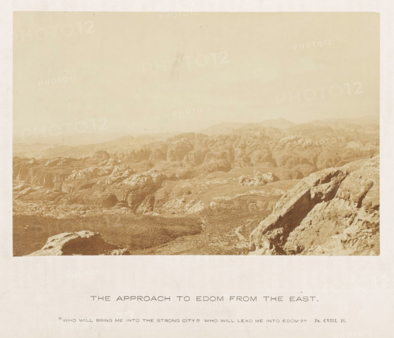 View of a landscape with mountains, referred to as Edom, The approach to Edom from the east, photograph, page, inscription: ''Who will bring me into the strong city? Who will lead me into Edom?' Ps. CVIII.10.', photographer: Francis Frith (possibly), c. 1850 - c. 1865, paper, albumen print, albumendruk, height 105 mm x width 157 mm, mountains, A sepia-toned photograph of a rocky, desert-like landscape with eroded rock formations and rugged hills extending toward a hazy horizon, with a foreground of large weathered boulders and a sloping rocky ridge on the right, and a wide pale sky occupying the upper portion of the image.