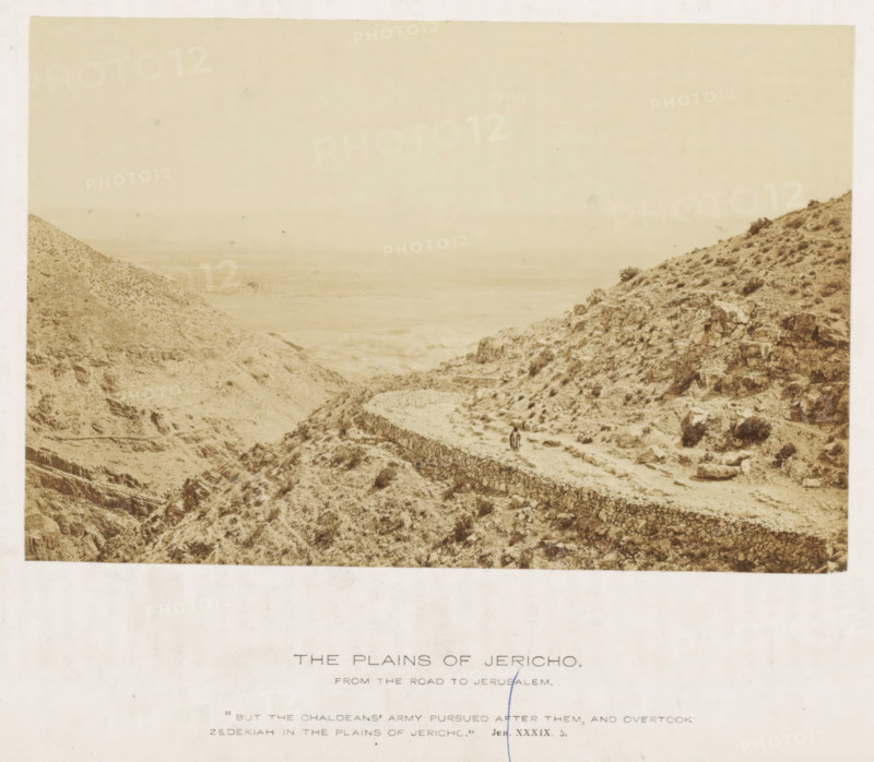 The plains of Jericho. From the road to Jerusalem, Landscape near Jericho, photograph, page, inscription: ''But the Chaldeans' army followed after them, and overtook Zedekiah in the plains of Jericho.' Jer XXXiX. 5.', photographer: Francis Frith (possibly), Jericho, c. 1850 - c. 1865, Francis Frith, photographer, Palestine, West Bank, paper, albumen print, height 104 mm x width 157 mm, mountains, desert - KK - ideal landscapes, A sepia-toned landscape photograph shows a rocky, arid terrain with steep hillsides and scattered low vegetation, a winding stone wall or terrace cutting across the slope toward the foreground, a narrow dirt path running alongside the wall, and two small human figures standing on the path; the background fades into a hazy plain and distant horizon under a pale sky.